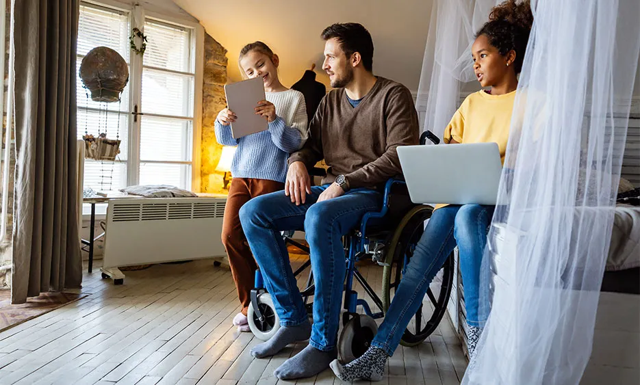 Familie sitzt gemeinsam in einem Wohnraum, ein Mann im Rollstuhl nutzt digitale Geräte mit zwei Kindern. - Deutsch Marketing