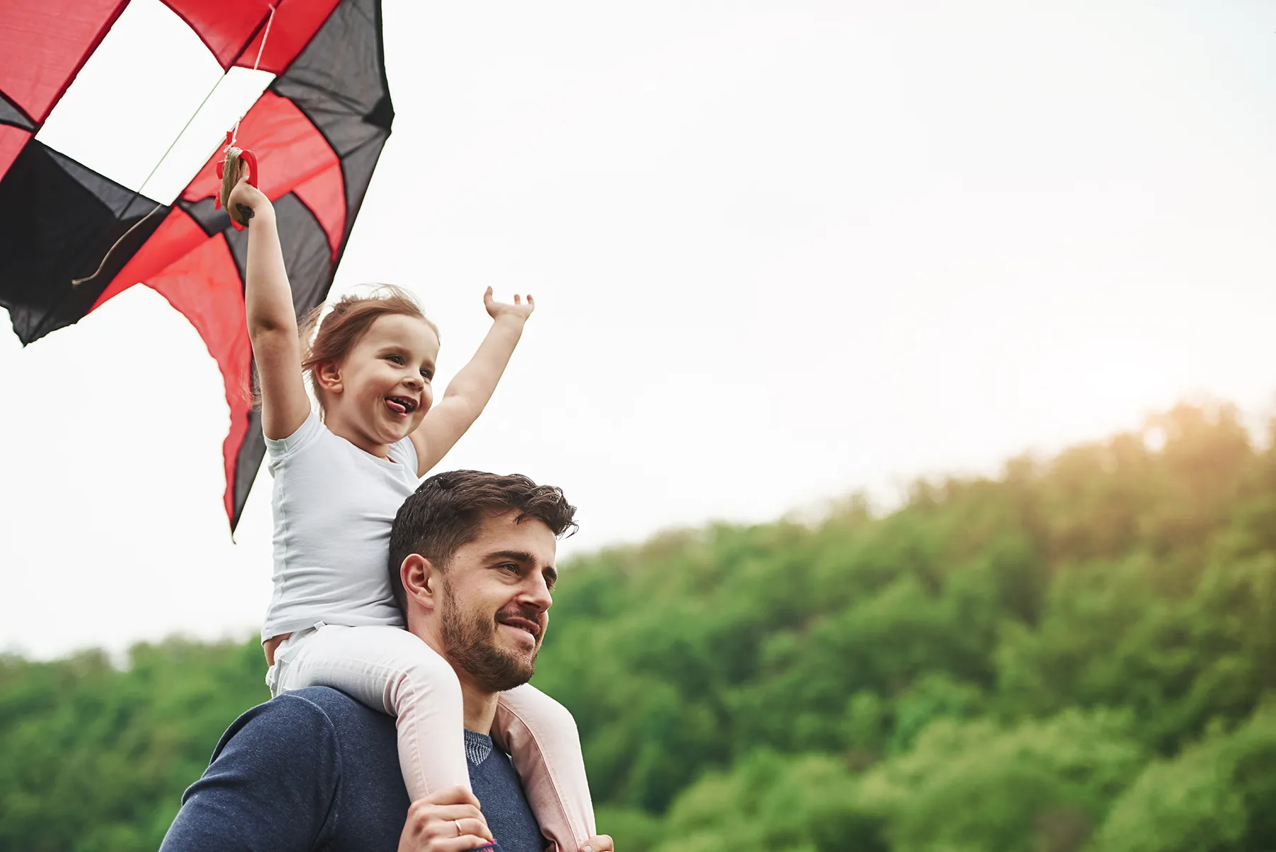 Vater trägt seine Tochter auf den Schultern, während sie fröhlich einen Drachen in der Natur hält. - Lebensversicherung Leads - Deutsch Marketing