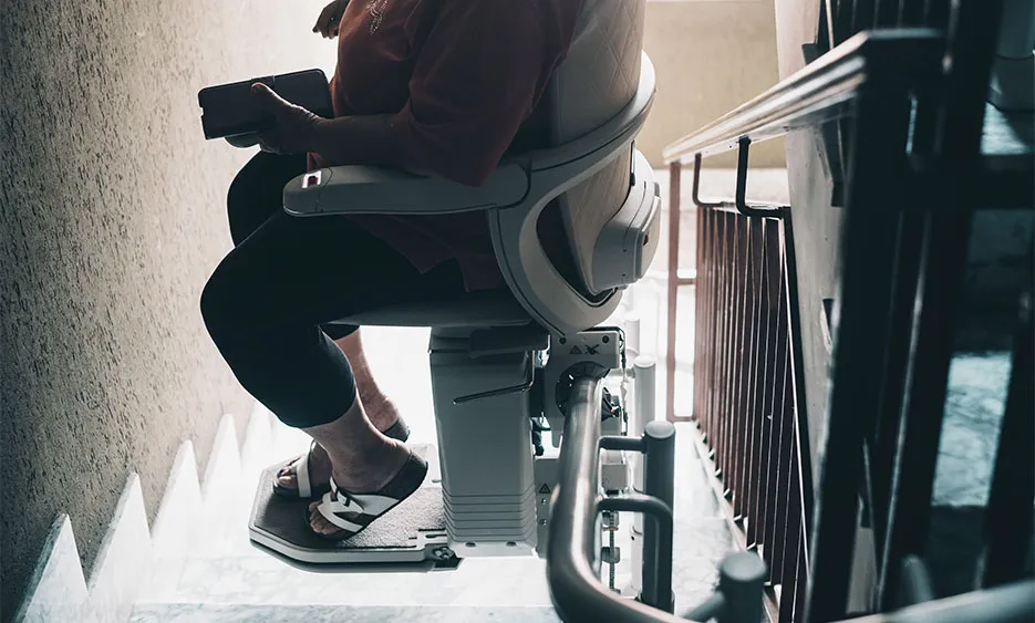 Person nutzt einen Treppenlift im Wohnhaus, während sie sitzend sicher eine Treppe hinauf- oder hinabfährt. - Deutsch Marketing GmbH
