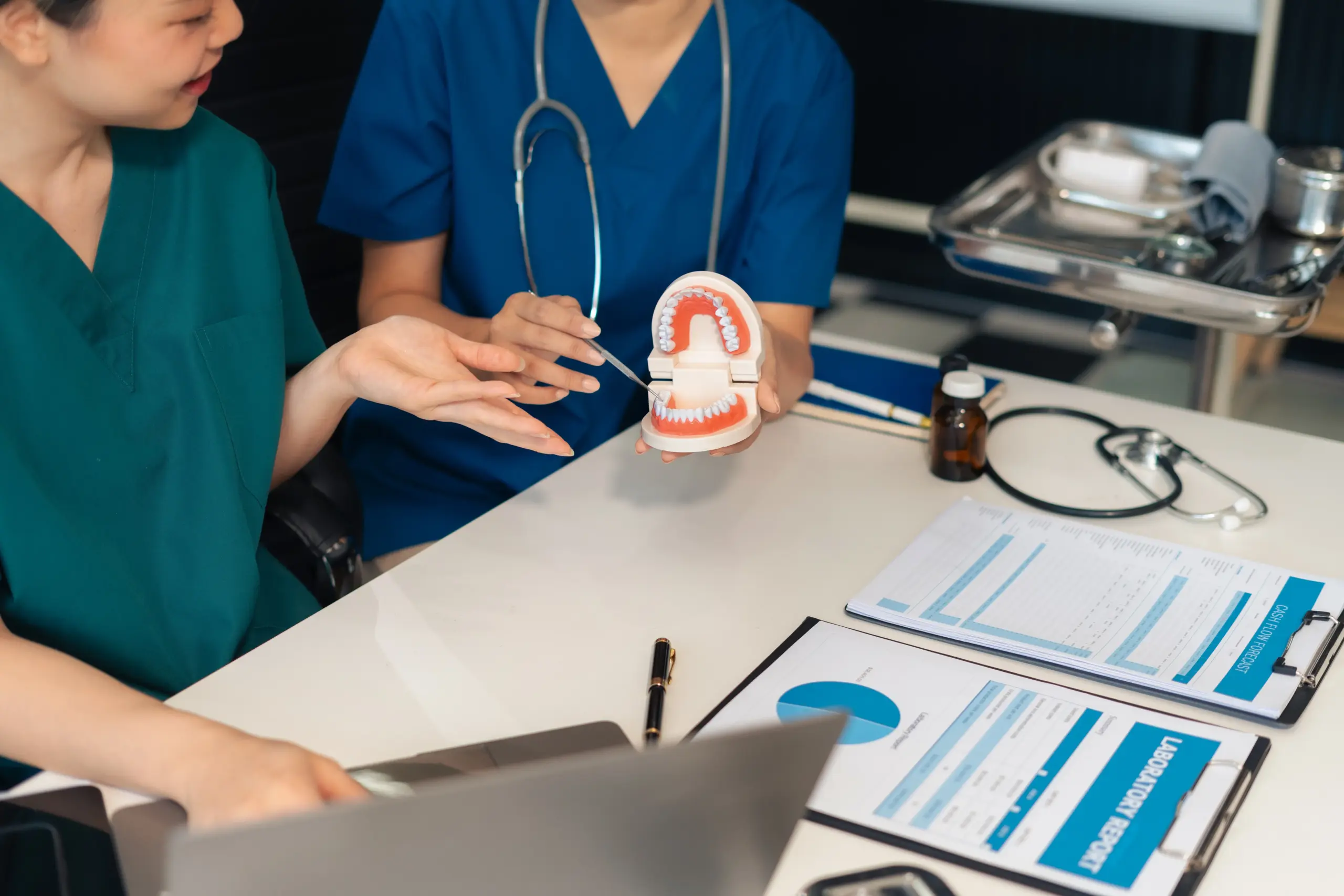 Zahnmedizinisches Fachpersonal erklärt an einem Gebissmodell eine Behandlung, während Unterlagen und ein Laptop auf dem Tisch liegen. - Deutsch Marketing