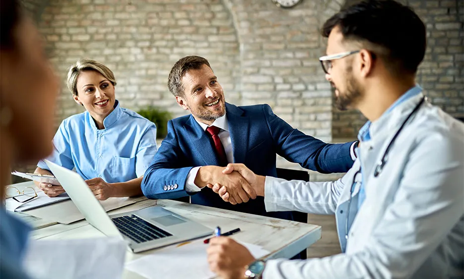 Beratungsgespräch zwischen Versicherungsberater und Ärzten mit Händedruck am Tisch, Laptop und Unterlagen liegen bereit. - Deutsch Marketing GmbH